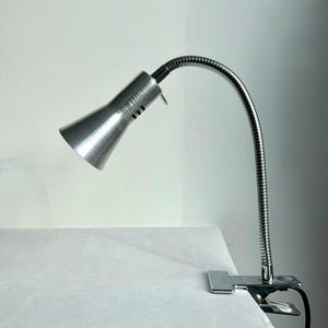 Könnte beinhalten: Eine silberfarbene Metall-Klemmlampe mit gebürsteter Oberfläche am konischen Schirm, einem flexiblen Schwanenhalsarm und einem stabilen Federklemmsockel. Die Lampe verfügt über ein schwarzes Stromkabel, das aus der Unterseite der Klemme herausführt.