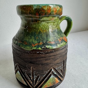 K&ouml;nnte beinhalten: Eine Keramikvase mit Henkel, mit einem strukturierten braunen Sockel mit geschnitzten geometrischen Mustern. Der obere Teil ist ein Farbverlauf aus Gr&uuml;n, Orange und Braun. Die Vase ist etwa 20 cm hoch.