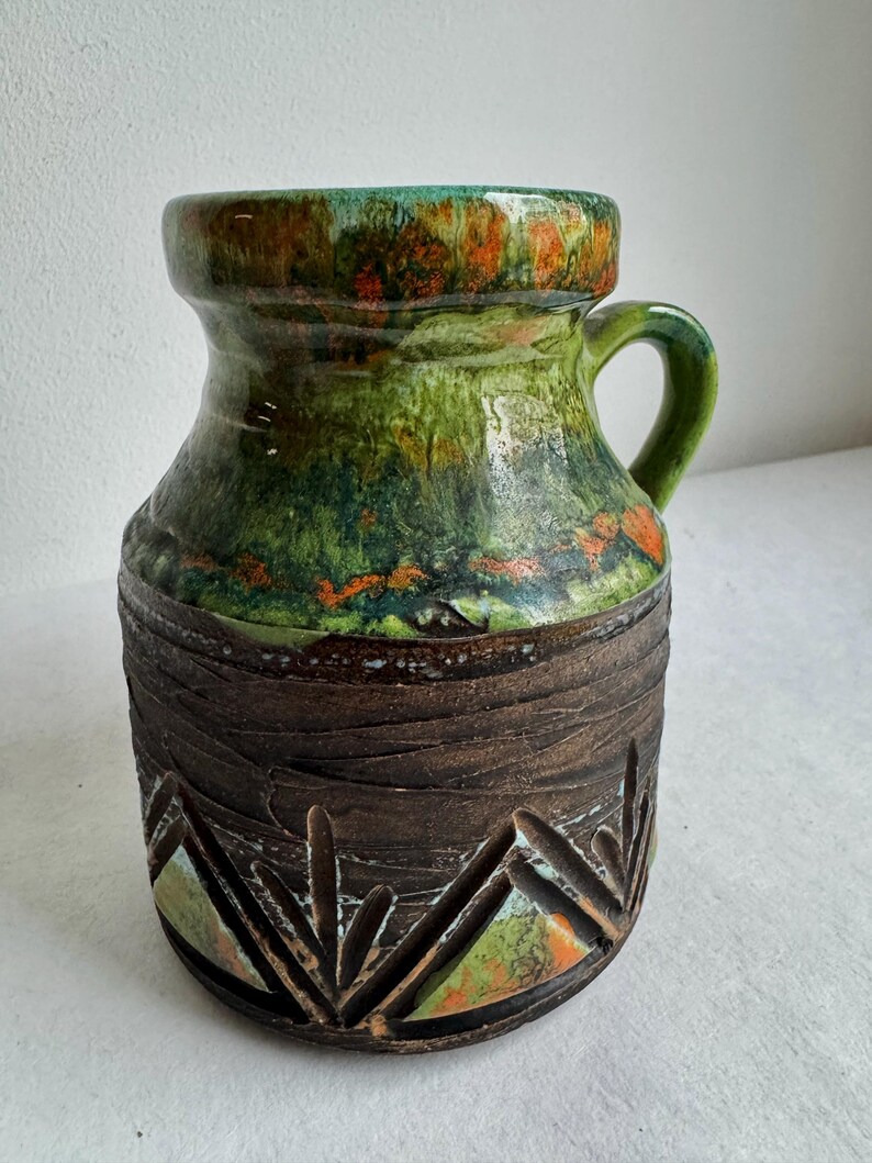 K&ouml;nnte beinhalten: Eine Keramikvase mit Henkel, mit einem strukturierten braunen Sockel mit geschnitzten geometrischen Mustern. Der obere Teil ist ein Farbverlauf aus Gr&uuml;n, Orange und Braun. Die Vase ist etwa 20 cm hoch.