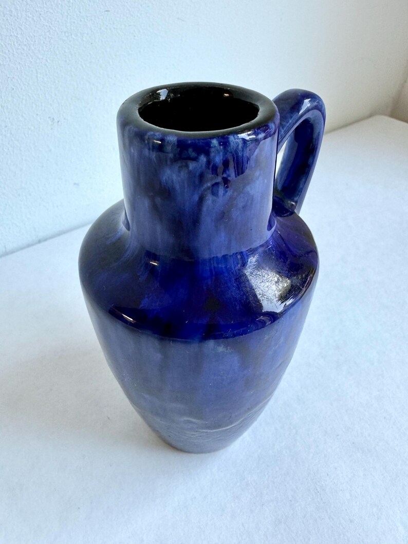 May include: A blue ceramic vase with a handle. The vase has a mottled glaze with a dark blue base and lighter blue streaks.