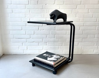 May include: A black metal side table with a glass top and a black bull sculpture on top. The table has a lower shelf with two books on it. The books are "Annie Leibovitz" and "Robert Frank, The Americans".