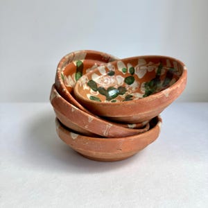 May include: A stack of four small, terracotta-colored bowls. The bowls have a shallow, rounded shape with a slightly flared rim. The interior of the top bowl features a floral pattern in green and white. The bowls appear to be handmade and have a rustic aesthetic.