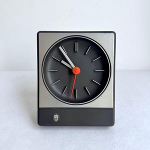 Könnte beinhalten: Eine quadratische Uhr im Retro-Stil mit schwarzem Zifferblatt, silbernem Rahmen und schwarzem Sockel. Die Uhr hat weiße Zeiger, einen roten Sekundenzeiger und einen orangefarbenen Punkt in der Mitte. Die Uhr ist etwa 10 cm breit und hoch.