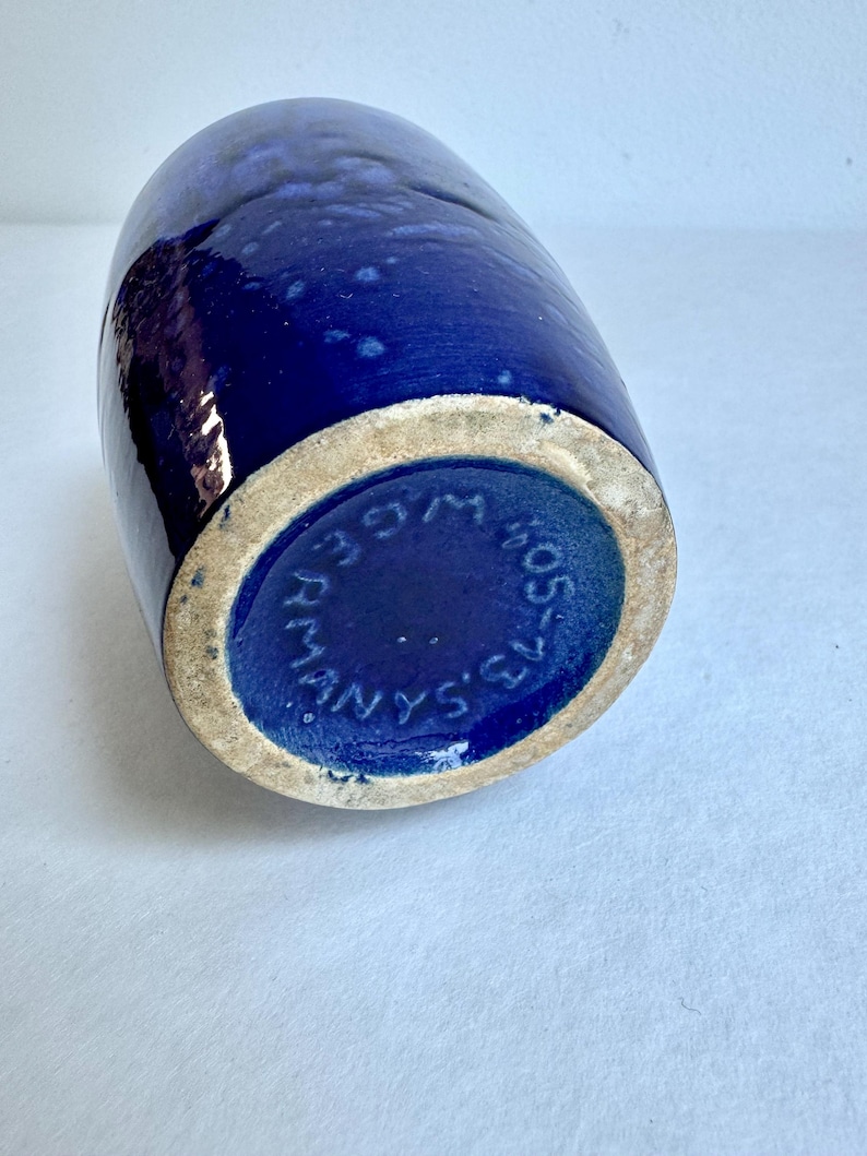 May include: A dark blue ceramic vase with a white rim and the text "MANYS'S" in a circle on the bottom.