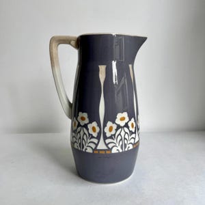 Op de afbeelding: Een keramische kan met een donkergrijze glazuurlaag en een herhalend patroon van witte bloemen met gele harten. De kan heeft een hoog, taps toelopend lichaam, een brede schenktuit en een gebogen handvat dat verloopt van grijs naar beige.