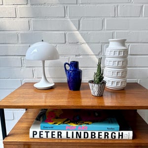 May include: A wooden side table with a black metal frame and a drawer. On the table are a white ceramic lamp with a white shade, a blue ceramic vase, a small potted succulent, and a white ceramic vase with a decorative pattern. On the shelf below are two books, one with a colorful cover and the title "POP ART" and the other with a white cover and the title "PETER LINDBERGH".