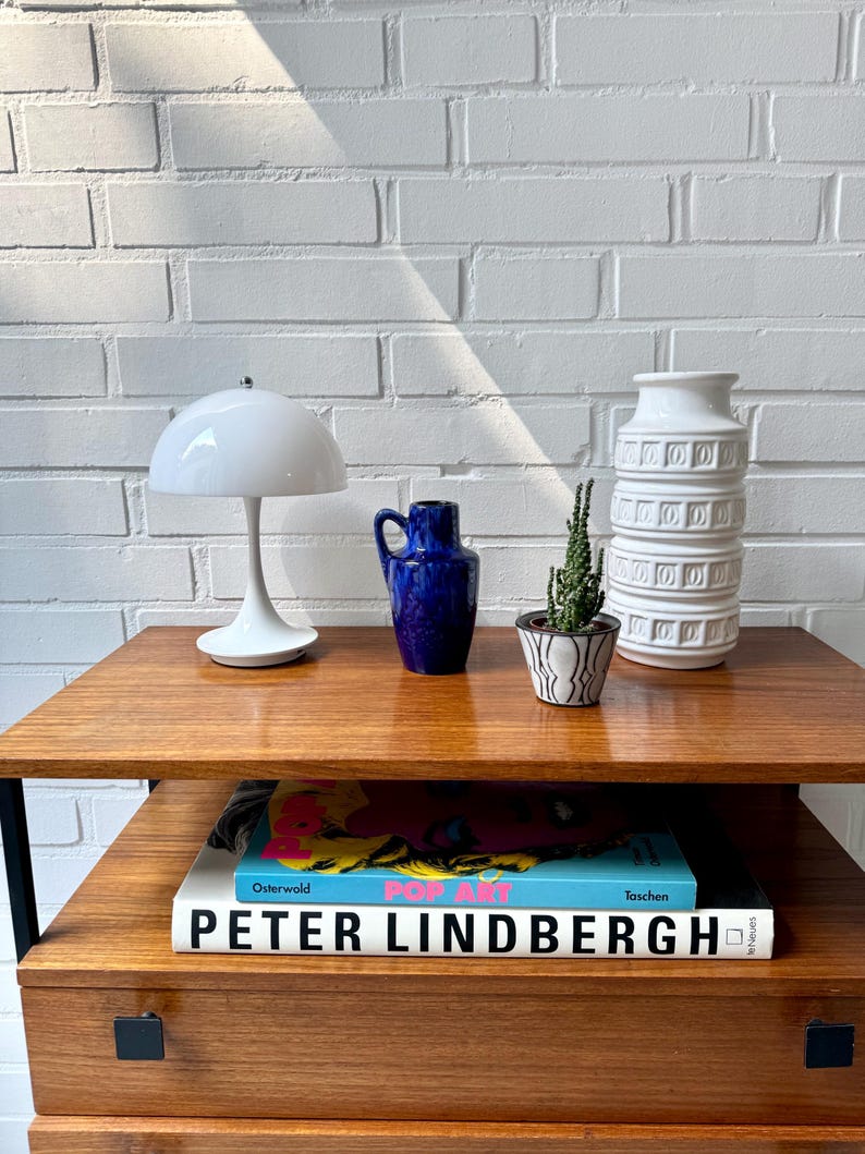 May include: A wooden side table with a black metal frame and a drawer. On the table are a white ceramic lamp with a white shade, a blue ceramic vase, a small potted succulent, and a white ceramic vase with a decorative pattern. On the shelf below are two books, one with a colorful cover and the title "POP ART" and the other with a white cover and the title "PETER LINDBERGH".