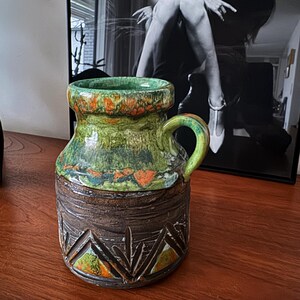 K&ouml;nnte beinhalten: Eine Vintage-Keramiktasse in Gr&uuml;n, Orange und Braun. Der obere Teil ist gr&uuml;n mit orangefarbenen Akzenten, der mittlere Teil ist strukturiertes Braun und der Boden hat ein geschnitztes geometrisches Muster. Sie hat einen gebogenen Griff.