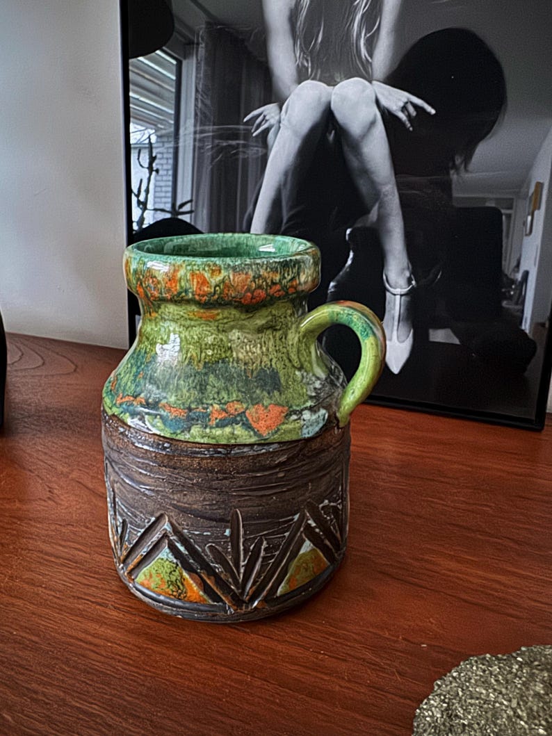 K&ouml;nnte beinhalten: Eine Vintage-Keramiktasse in Gr&uuml;n, Orange und Braun. Der obere Teil ist gr&uuml;n mit orangefarbenen Akzenten, der mittlere Teil ist strukturiertes Braun und der Boden hat ein geschnitztes geometrisches Muster. Sie hat einen gebogenen Griff.