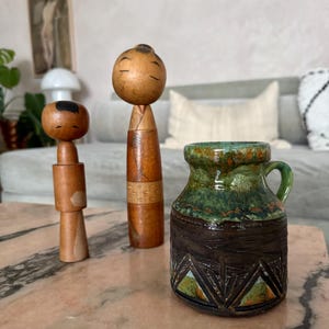 K&ouml;nnte beinhalten: Drei dekorative Gegenst&auml;nde auf einer rosa Marmoroberfl&auml;che. Zwei Kokeshi-Puppen aus Holz mit minimalistisch bemalten Gesichtern stehen neben einem kleinen, strukturierten Keramikkrug mit gr&uuml;ner und brauner Glasur. Der Krug hat einen kleinen Griff.