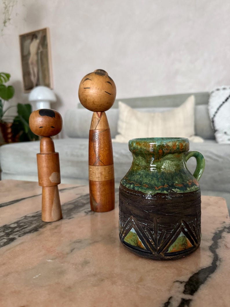 K&ouml;nnte beinhalten: Drei dekorative Gegenst&auml;nde auf einer rosa Marmoroberfl&auml;che. Zwei Kokeshi-Puppen aus Holz mit minimalistisch bemalten Gesichtern stehen neben einem kleinen, strukturierten Keramikkrug mit gr&uuml;ner und brauner Glasur. Der Krug hat einen kleinen Griff.
