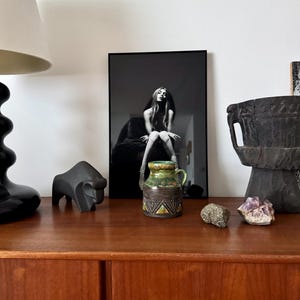 K&ouml;nnte beinhalten: Ein Holzschrank mit verschiedenen dekorativen Gegenst&auml;nden. Ein schwarz-wei&szlig; gerahmtes Foto, eine schwarze Lampe, eine schwarze Tierskulptur, eine kleine Keramikvase in Gr&uuml;n- und Braunt&ouml;nen, ein Stein und ein lila Kristall.