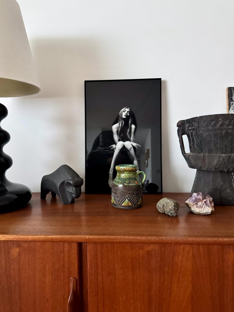 K&ouml;nnte beinhalten: Ein Holzschrank mit verschiedenen dekorativen Gegenst&auml;nden. Ein schwarz-wei&szlig; gerahmtes Foto, eine schwarze Lampe, eine schwarze Tierskulptur, eine kleine Keramikvase in Gr&uuml;n- und Braunt&ouml;nen, ein Stein und ein lila Kristall.