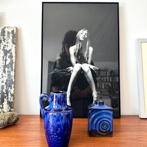 May include: Two blue ceramic vases, one with a round body and a handle, the other with a square body and a swirling pattern. The vases are on a wooden surface in front of a black and white photo.