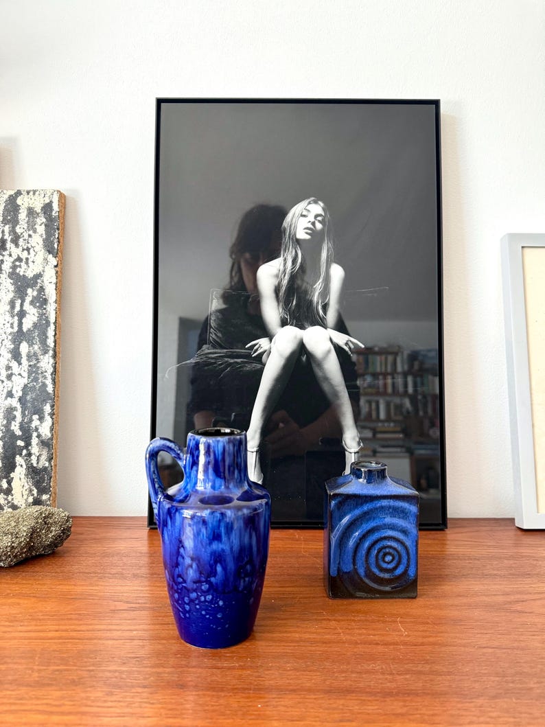 May include: Two blue ceramic vases, one with a round body and a handle, the other with a square body and a swirling pattern. The vases are on a wooden surface in front of a black and white photo.
