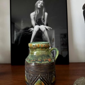 K&ouml;nnte beinhalten: Kleine, handgefertigte Keramikvase mit Henkel. Die Vase hat einen dunkelbraunen Sockel mit geometrischen Schnitzereien und einen gr&uuml;n-orangen Rand. Im Hintergrund befindet sich eine Schwarz-Wei&szlig;-Fotografie einer Frau.