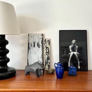 May include: A wooden table with a black lamp, a black ceramic bison sculpture, two wooden boards, a blue and white ceramic vase, a blue and white ceramic box, and a framed black and white photograph of a woman sitting on a bench.
