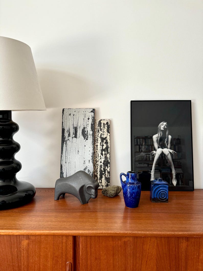 May include: A wooden table with a black lamp, a black ceramic bison sculpture, two wooden boards, a blue and white ceramic vase, a blue and white ceramic box, and a framed black and white photograph of a woman sitting on a bench.
