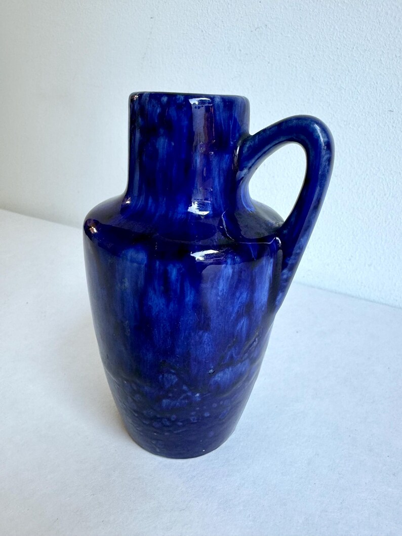 May include: A cobalt blue ceramic vase with a single handle. The vase has a mottled glaze with a slightly textured surface.