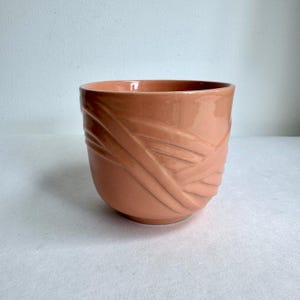 Pode incluir: Um vaso de cerâmica cor de pêssego com acabamento brilhante. O vaso apresenta um design envolvente e texturizado de linhas diagonais. A forma arredondada e a cor quente tornam-no adequado para várias plantas e decoração de casa.