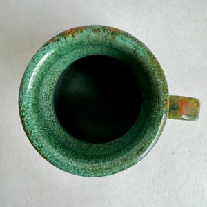 K&ouml;nnte beinhalten: Eine handgefertigte Keramiktasse mit gl&auml;nzender gr&uuml;ner Glasur. Die Tasse hat einen abgerundeten K&ouml;rper, einen kleinen Henkel und einen dunklen Innenraum. Der Rand hat ein strukturiertes Aussehen mit Hauch von Orange und Braun.