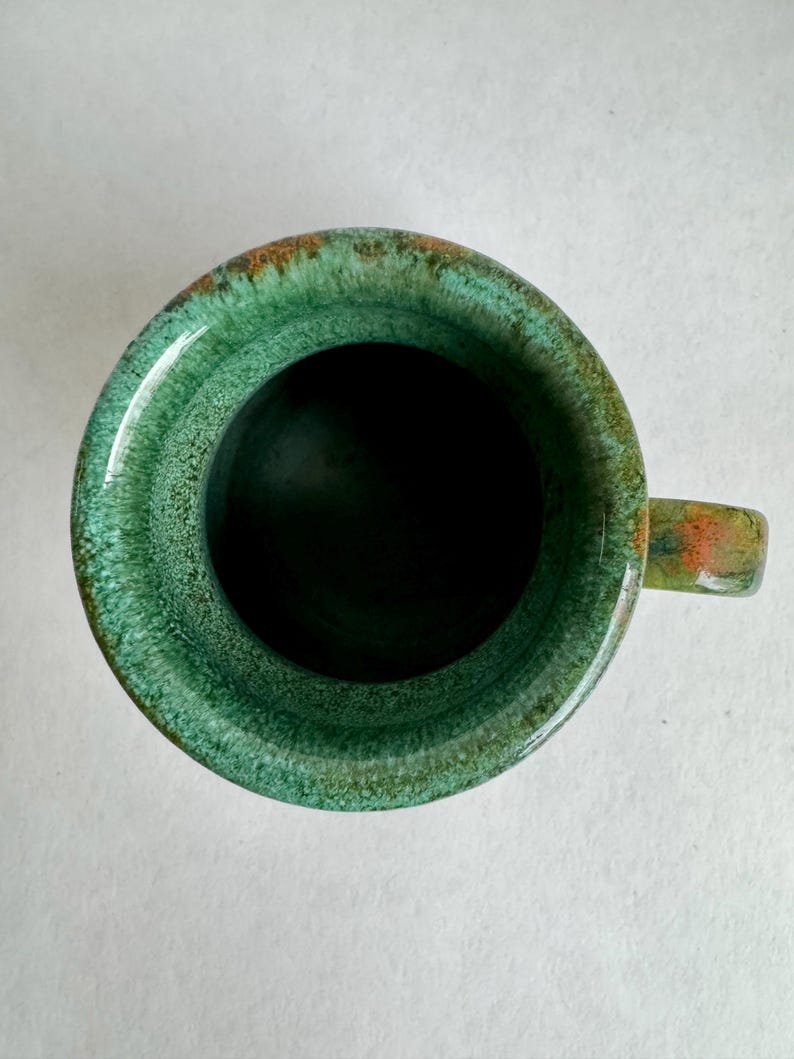 K&ouml;nnte beinhalten: Eine handgefertigte Keramiktasse mit gl&auml;nzender gr&uuml;ner Glasur. Die Tasse hat einen abgerundeten K&ouml;rper, einen kleinen Henkel und einen dunklen Innenraum. Der Rand hat ein strukturiertes Aussehen mit Hauch von Orange und Braun.