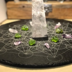 Puede incluir: Una placa de pizarra negra con un diseño geométrico grabado. Un cristal de cuarzo transparente se encuentra en el centro de la placa, rodeado de cristales verdes y morados.