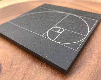 May include: A black slate coaster with a white engraved design of the Fibonacci spiral, also known as the Golden Spiral.