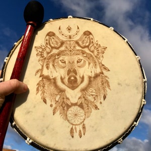 May include: A brown leather drum with a black rim and a wolf design. The drum is being held by a hand with a wooden stick. The background is a blue sky with white clouds.