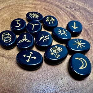 May include: A collection of thirteen smooth, oval-shaped black stones, each featuring a unique gold-colored engraved symbol. The symbols include an eye, a crescent moon, a sun, arrows, and various abstract geometric patterns etched into the surface.