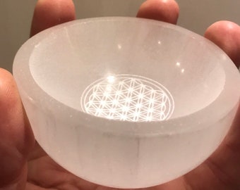 May include: A white selenite bowl with a flower of life design etched into the bottom. The bowl is smooth and polished.