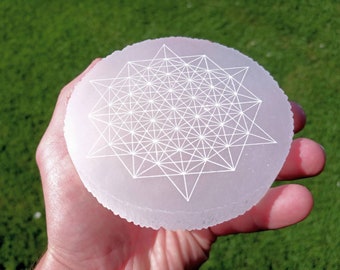 May include: A round, pale pink selenite crystal with a white geometric pattern etched on its surface. The crystal is held in a hand against a backdrop of green grass. The design features intersecting lines and star shapes.