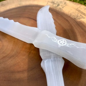 May include: Two white selenite crystal wands, one with a pentagram design. The wands are on a wooden surface.