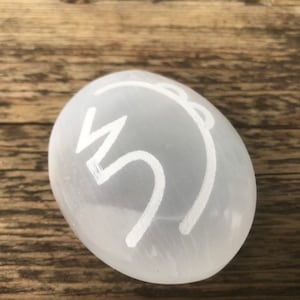 Pode incluir: Ovo de cristal de selenita branco com um s&iacute;mbolo branco desenhado nele. O ovo est&aacute; sobre uma superf&iacute;cie de madeira.