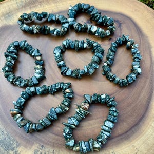Könnte beinhalten: Sieben Armbänder aus kleinen, unregelmäßig geformten grünen und grauen Steinen. Die Armbänder sind auf einer runden Holzoberfläche mit natürlicher Maserung angeordnet. Die Steine haben ein gesprenkeltes Aussehen, und die Armbänder sind kreisförmig.