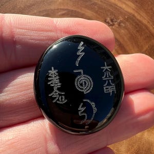 以下が含まれることがあります： 刻まれたシンボルが付いた黒い楕円形の石。石は光沢のある仕上げで、人の手で持たれています。シンボルは銀色で、アジア起源のようです。石は滑らかで磨かれています。