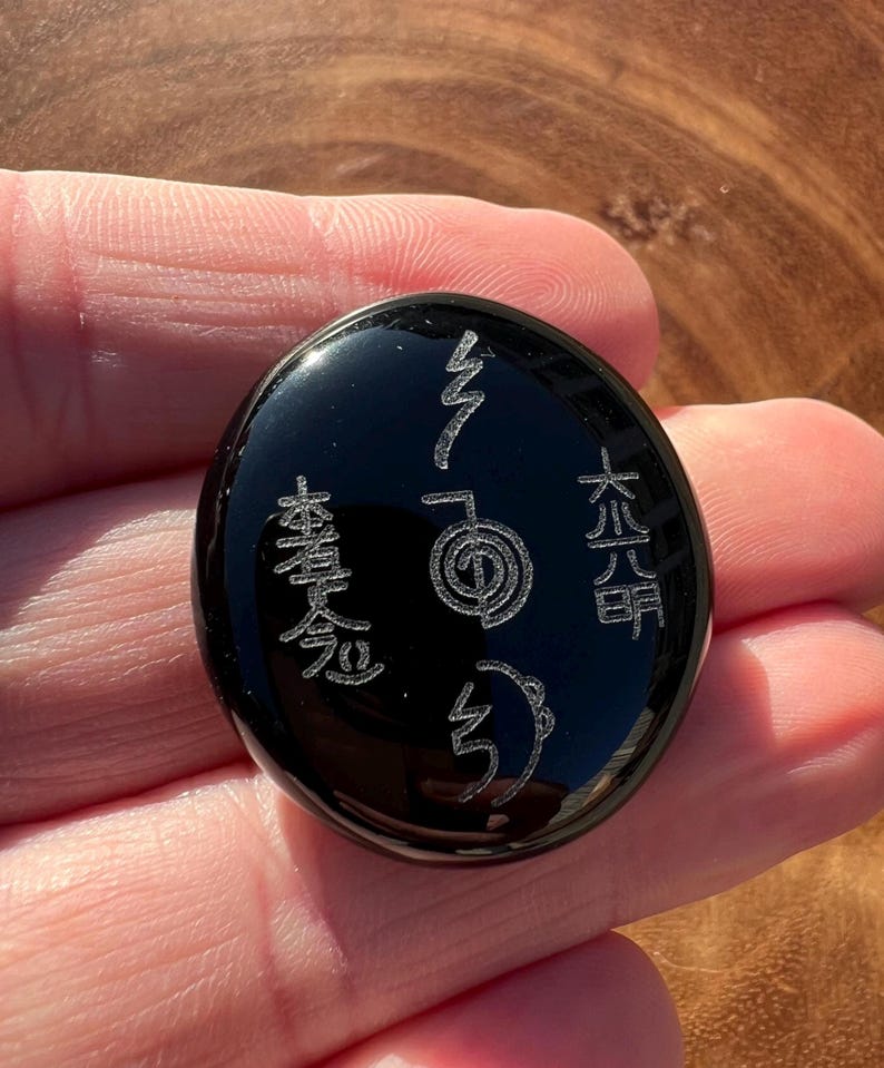 以下が含まれることがあります： 刻まれたシンボルが付いた黒い楕円形の石。石は光沢のある仕上げで、人の手で持たれています。シンボルは銀色で、アジア起源のようです。石は滑らかで磨かれています。
