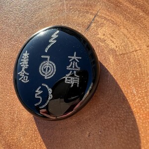 以下が含まれることがあります： 黒い丸い石に、エッチングされたシンボルと日本語の文字が刻まれています。研磨された表面は光を反射し、銀色のデザインを際立たせています。この石は、精神的なアイテムまたは装飾品です。