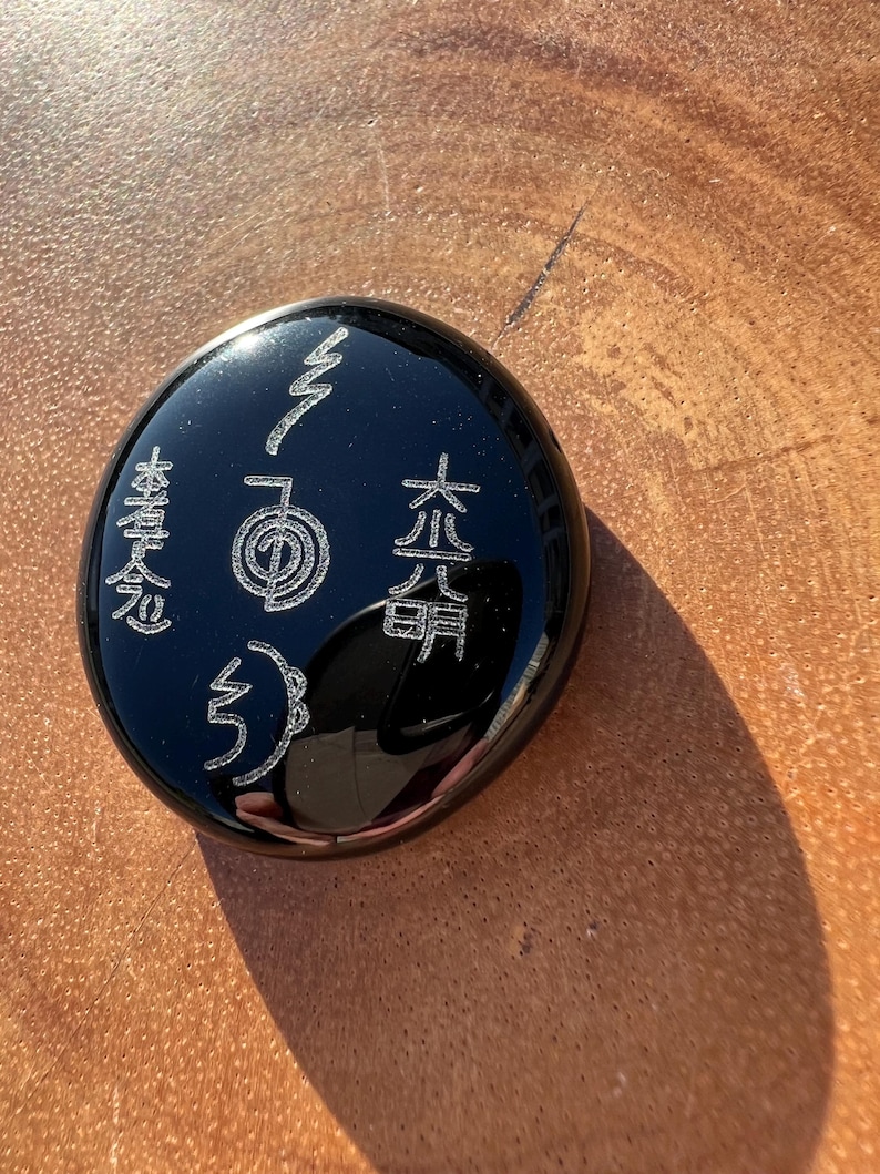 以下が含まれることがあります： 黒い丸い石に、エッチングされたシンボルと日本語の文字が刻まれています。研磨された表面は光を反射し、銀色のデザインを際立たせています。この石は、精神的なアイテムまたは装飾品です。
