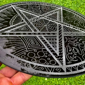 Peut inclure: Une assiette ronde en verre noir avec un motif de pentagramme argenté. Le pentagramme est divisé en cinq sections, chacune avec un motif complexe différent.