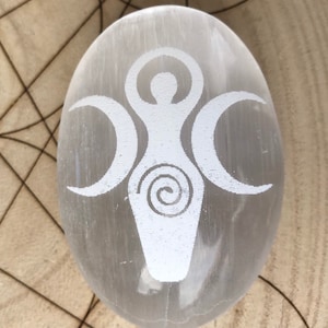 Peut inclure: Œuf en cristal de sélénite blanc avec un symbole de déesse triple lune gravé. Le symbole comprend une figure féminine avec les bras levés et trois croissants de lune.