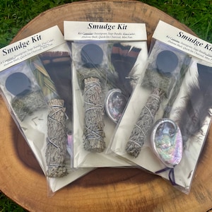 May include: Three smudge kits, each containing a bundle of sage, a small abalone shell, a piece of charcoal, and a feather fan. The kits are packaged in clear plastic bags with white labels that read "Smudge Kit" and list the kit contents.