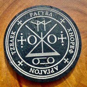 May include: Black circular slate coaster with a silver inscription and a silver symbol in the centre. The inscription reads "APLATON", "O.O", "X.HOPE.X", "TZBA.K", "RAGUEL", and "X".