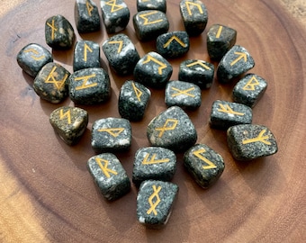 May include: A set of 24 black and white speckled stone runes with gold painted symbols. The runes are arranged on a wooden surface.