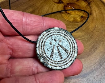 May include: A circular pendant necklace with a black cord. The pendant features a white and grey speckled background with a bronze-coloured circular design and three lines pointing downwards. The pendant is held by a hand.