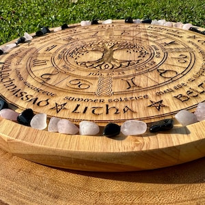 May include: A wooden circular board with a tree of life design and a calendar of the eight sabbats. The board is surrounded by a ring of pink and black stones.