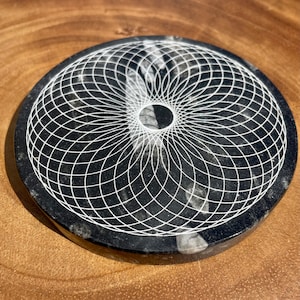 May include: A black and white round stone coaster with a geometric design carved into the surface. The design is a series of concentric circles that create a mandala-like pattern.