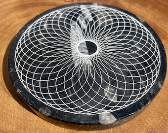 May include: A black and white round stone coaster with a geometric design carved into the surface. The design is a series of concentric circles that create a mandala-like pattern.