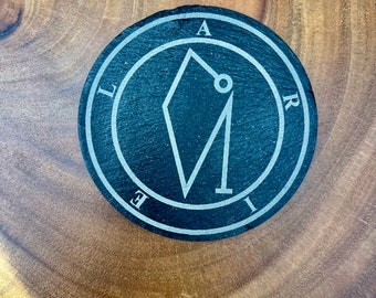 May include: A black slate coaster with a silver sigil inscribed in a circle. The sigil is a geometric shape with a circle in the center. The letters "ALIRE" are inscribed around the circle.