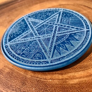 Pode incluir: Um descanso de copo de vidro azul com um design gravado branco de uma estrela de cinco pontas com padrões intrincados dentro de cada ponta.